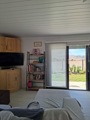 A bedroom with a bed, wooden cabinets, a bookshelf, a television, and a sliding glass door leading to a view of a field. - Relaxed 2-bedroom, family and small-pet-friendly cottage with farm views. (Moorreesburg)