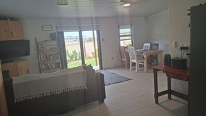 Una sala de estar con un sofá, un televisor, una mesa de comedor con sillas, un ventilador de techo y vistas por una ventana.