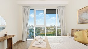 A hotel room with a large window, a bed with white linens, a wooden bench, and a painting on the wall. - 2501-Bloomfields Luxurious 3BR  West Res (Dubai)