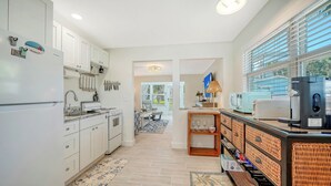 A modern kitchen with white cabinets, a stove, and a coffee machine. There is a dining area with a table and chairs, and a view of the outdoors through large windows.