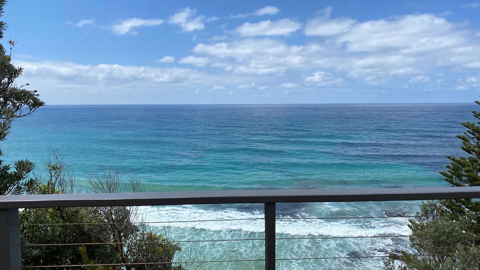 Blick auf das Meer von einem Balkon mit Geländer, Bäumen und blauem Himmel.