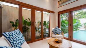Een moderne woonkamer met een bank, een rond houten bijzettafeltje en een stoel met een gekleurd kussen. Grote glazen deuren leiden naar een zwembadgedeelte met weelderige begroeiing.