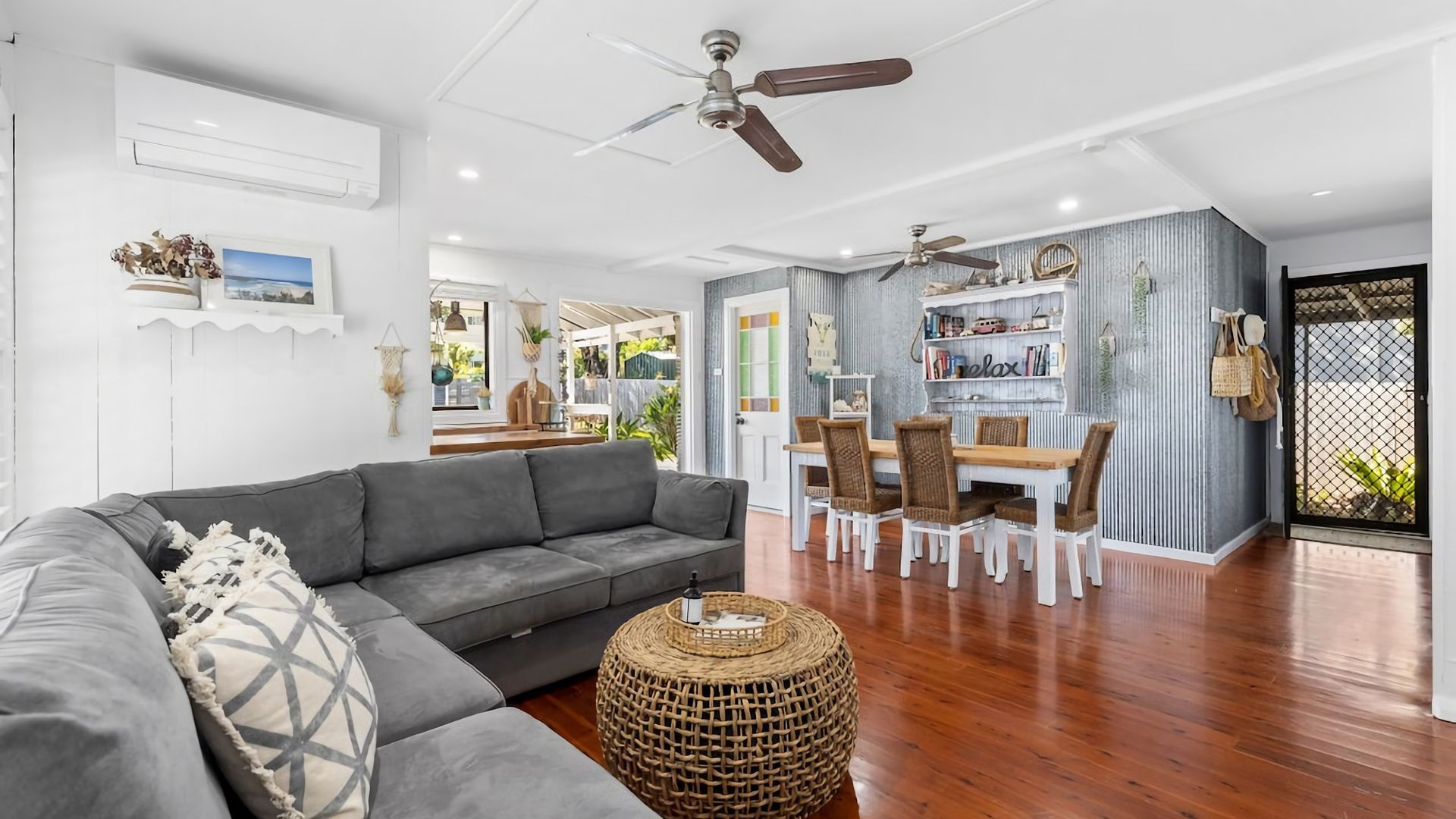 A modern living room with a grey sectional sofa, a round wicker ottoman, and a dining area with white chairs and a wooden table.