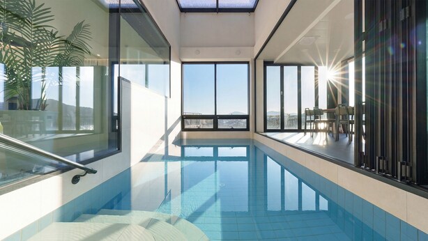 Une piscine intérieure avec des parois en verre transparent, offrant une vue sur l’extérieur, et un espace baigné de lumière naturelle avec des fauteuils.