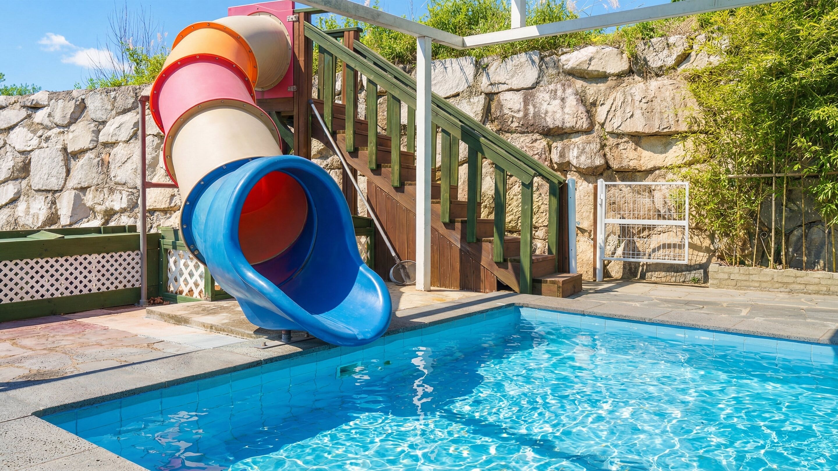 A swimming pool with a multi-colored slide, a stone wall, and a wooden deck.