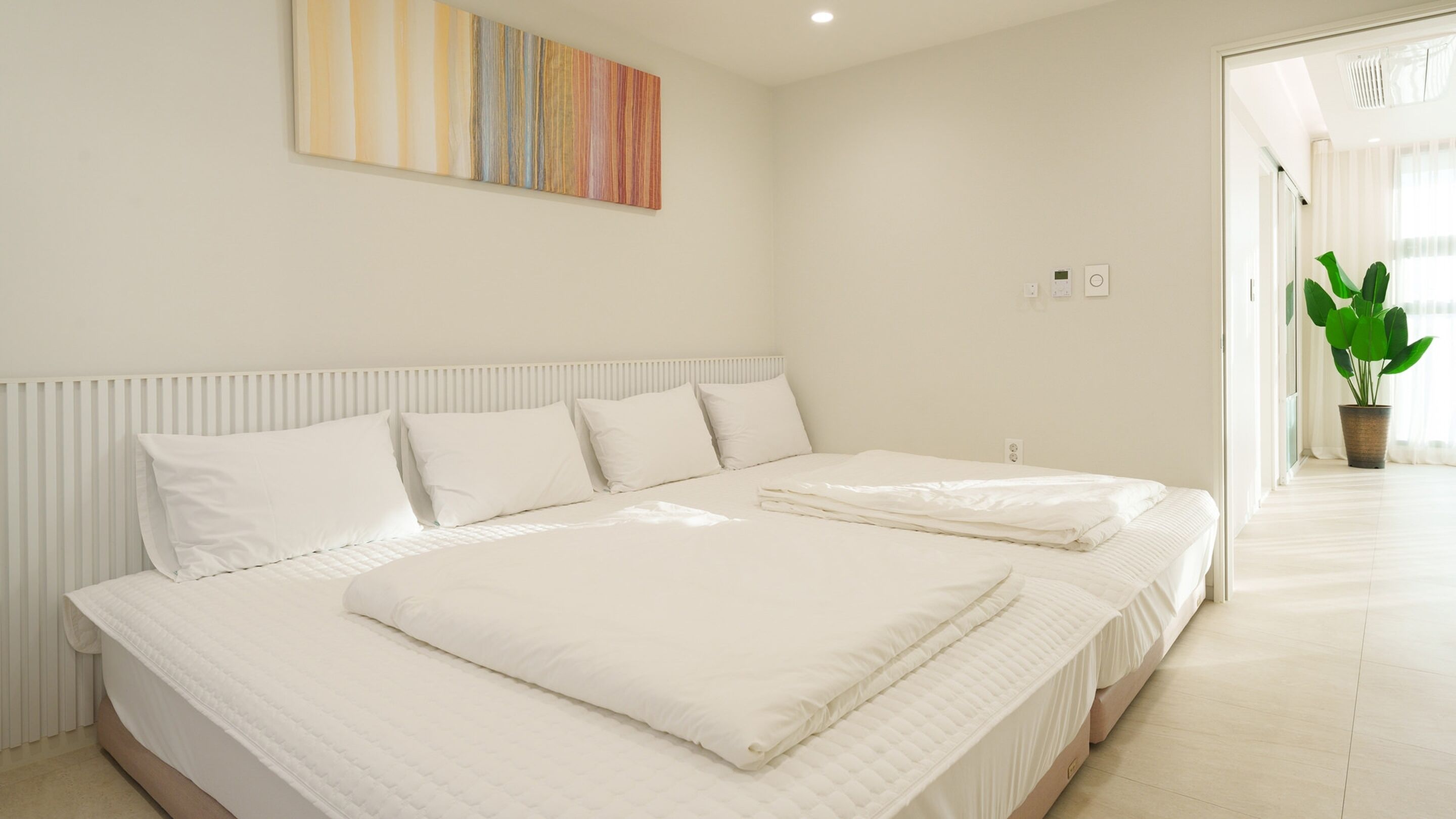 A modern bedroom with a large bed, white bedding, a radiator, a wall-mounted artwork, and a potted plant.