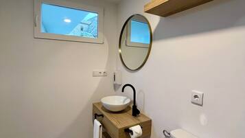 A bathroom with a window, a round mirror, a sink, a toilet, and a wooden shelf.