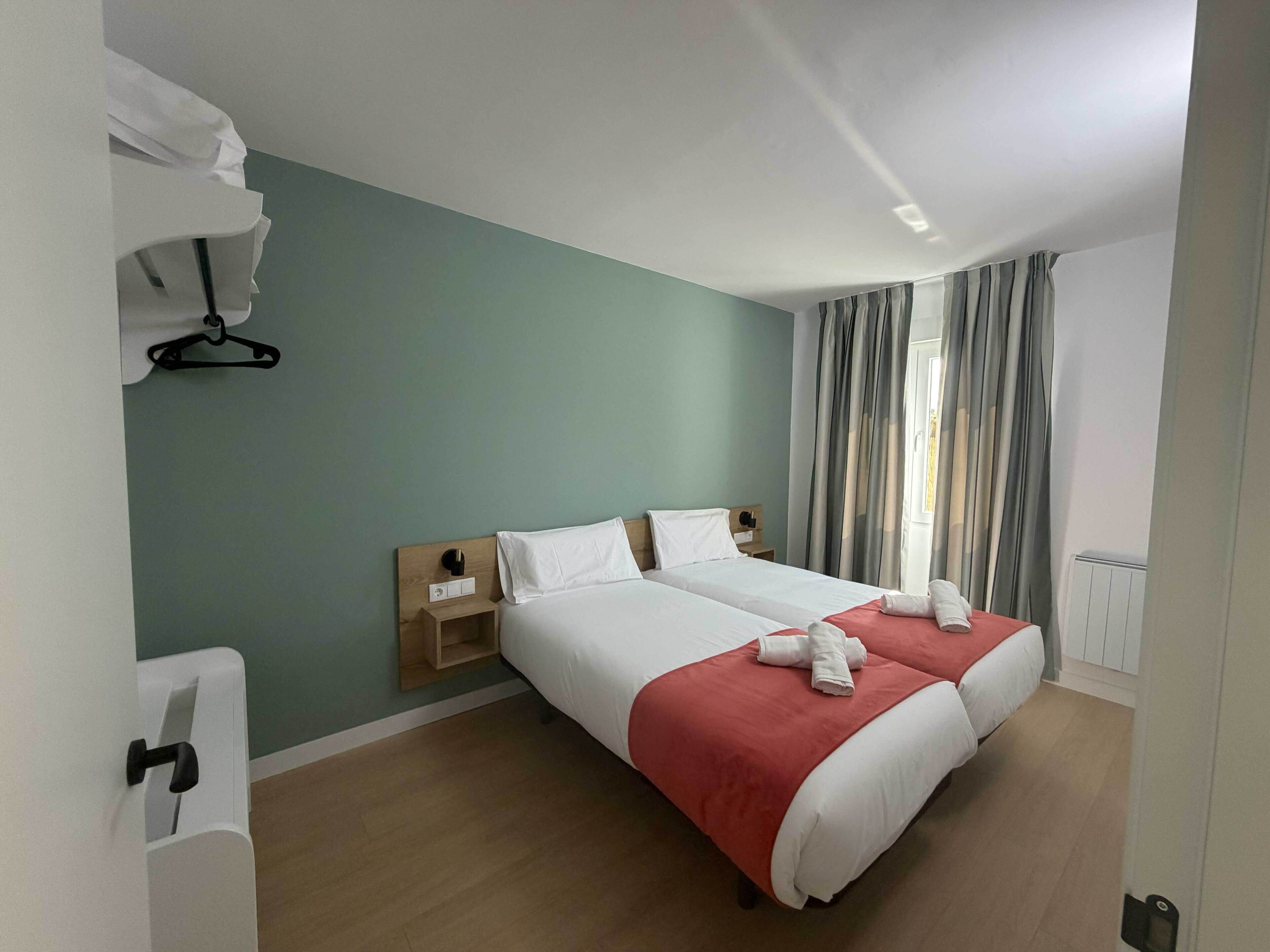 A hotel room with two beds, a wooden headboard, a nightstand, a window with curtains, and a radiator.