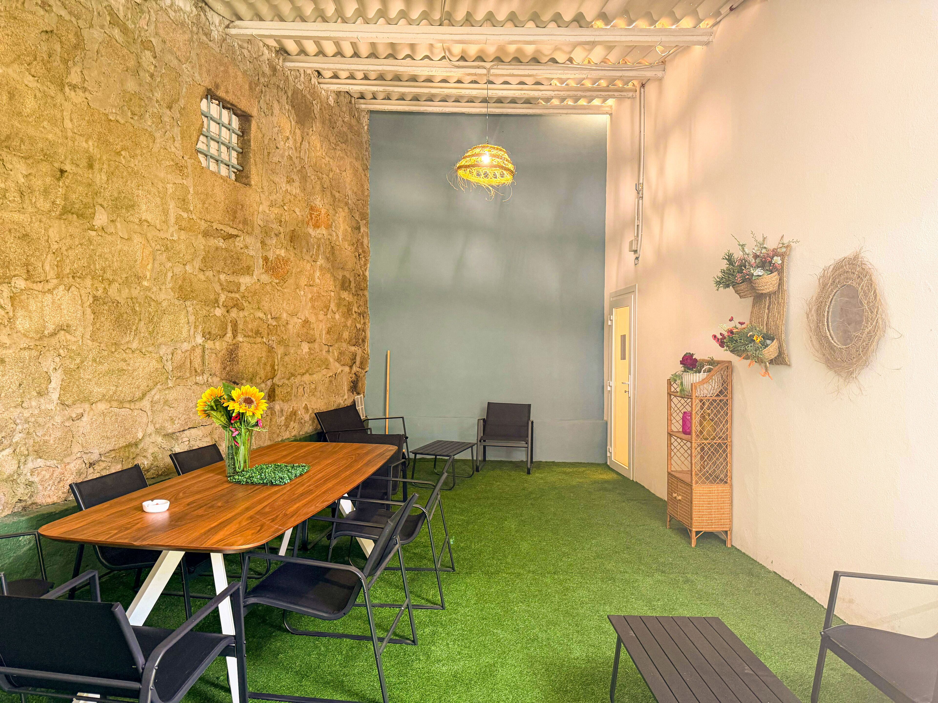 A room with a wooden table, black chairs, a green artificial turf floor, a stone wall, a hanging light fixture, and a door leading to another room.