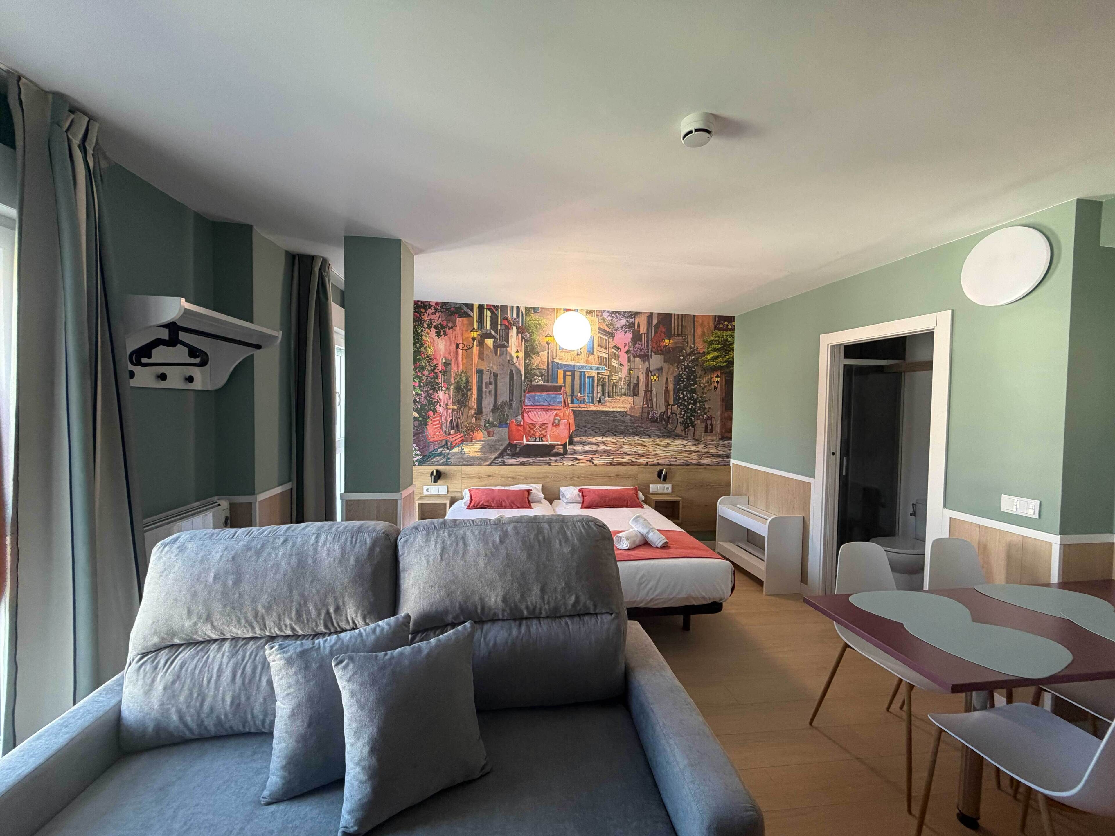 A modern hotel room with a sofa, a bed, a dining table, and a mural of a street scene.