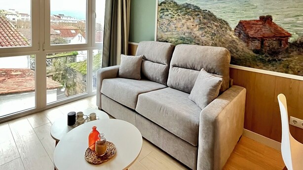 A living room with a grey sofa, a small round table with a drink, and a painting of a coastal scene. - Luxury Singular Apartments Padrón (Padron)