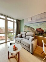 Un salón con un sofá gris, una mesita redonda con una bebida y un cuadro de una escena costera.