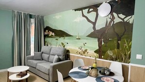 A room with a sofa, two dining tables, chairs, a mural of a beach scene, and a window with curtains. - Luxury Singular Apartments Padrón (Padron)