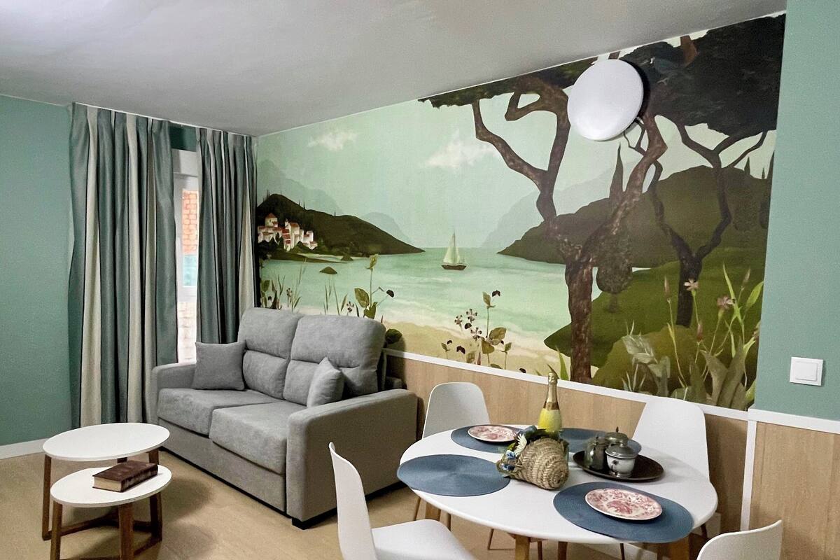 Une pièce comprenant un canapé, deux tables à manger, des chaises, une fresque représentant une scène de plage et une fenêtre avec des rideaux.