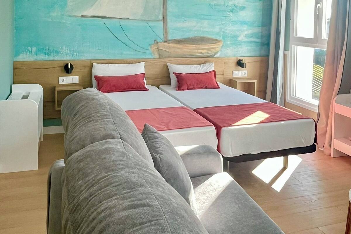 Une chambre d’hôtel avec un lit, un canapé, un tableau représentant un voilier et une fenêtre avec des rideaux.