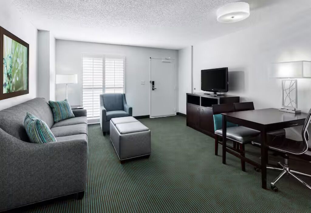 A modern hotel room with a grey sofa, a matching ottoman, a dining table with chairs, a flat-screen TV, and a lamp.