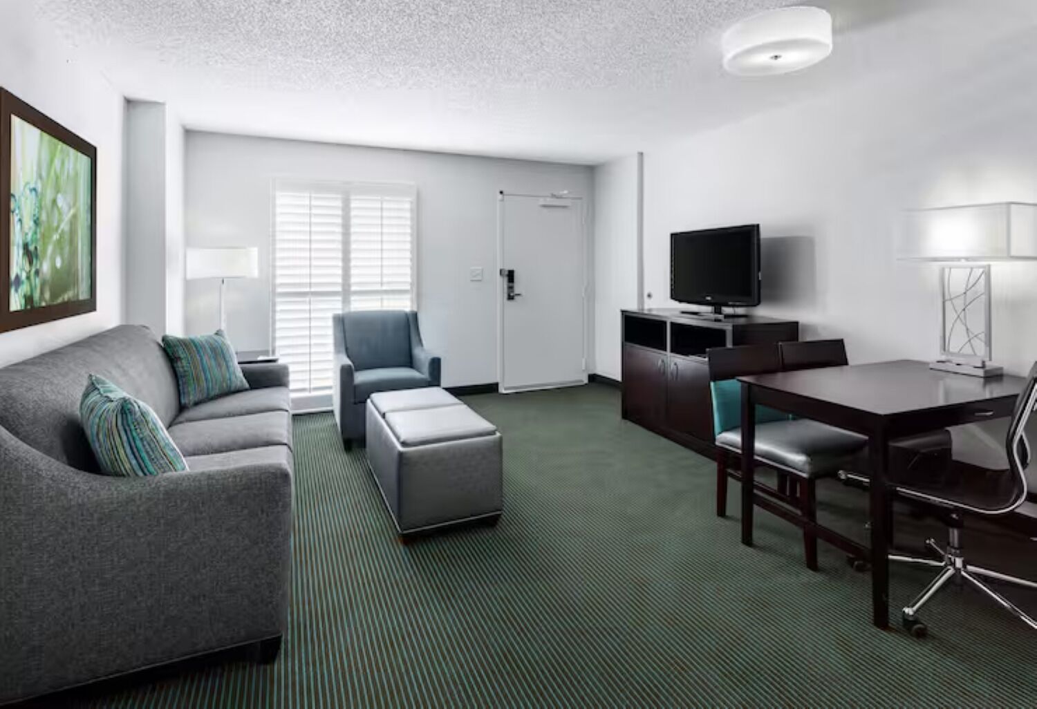 Een moderne hotelkamer met een grijze bank, een klein poufje, een eettafel met stoelen, een flatscreen televisie en een lamp.