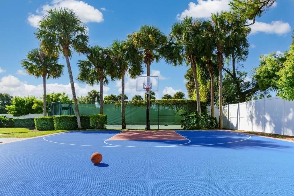 Un terrain de basket en plein air, doté d’une surface bleue, entouré de palmiers et délimité par une clôture blanche.