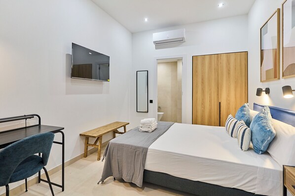 Une chambre à coucher moderne comprenant un lit, une table de chevet, une chaise, un téléviseur, une armoire et un petit banc.