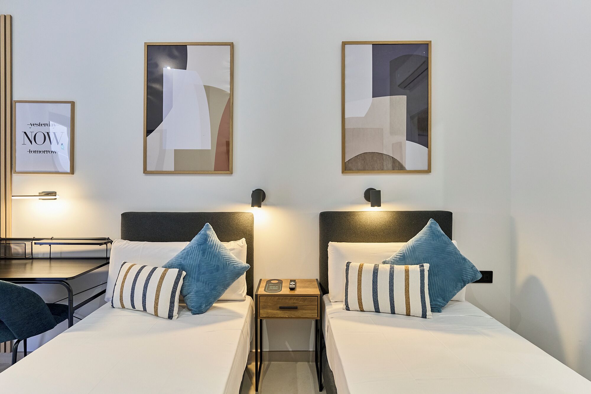 Una habitación de hotel con dos camas, cada una con una almohada y una mesita de noche con lámpara. En las paredes hay cuadros enmarcados y un letrero con las palabras 'yesterday' y 'now' a la izquierda.