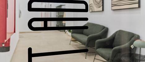 An interior space with a modern design, featuring a glass partition, green upholstered chairs, and abstract wall art.