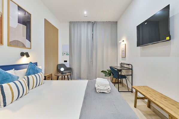 A modern hotel room with a neatly made bed, a desk with a chair, a bench, a TV mounted on the wall, and a large window with curtains. - Sant Adrià Urban Suites by gaiarooms (Badalona)