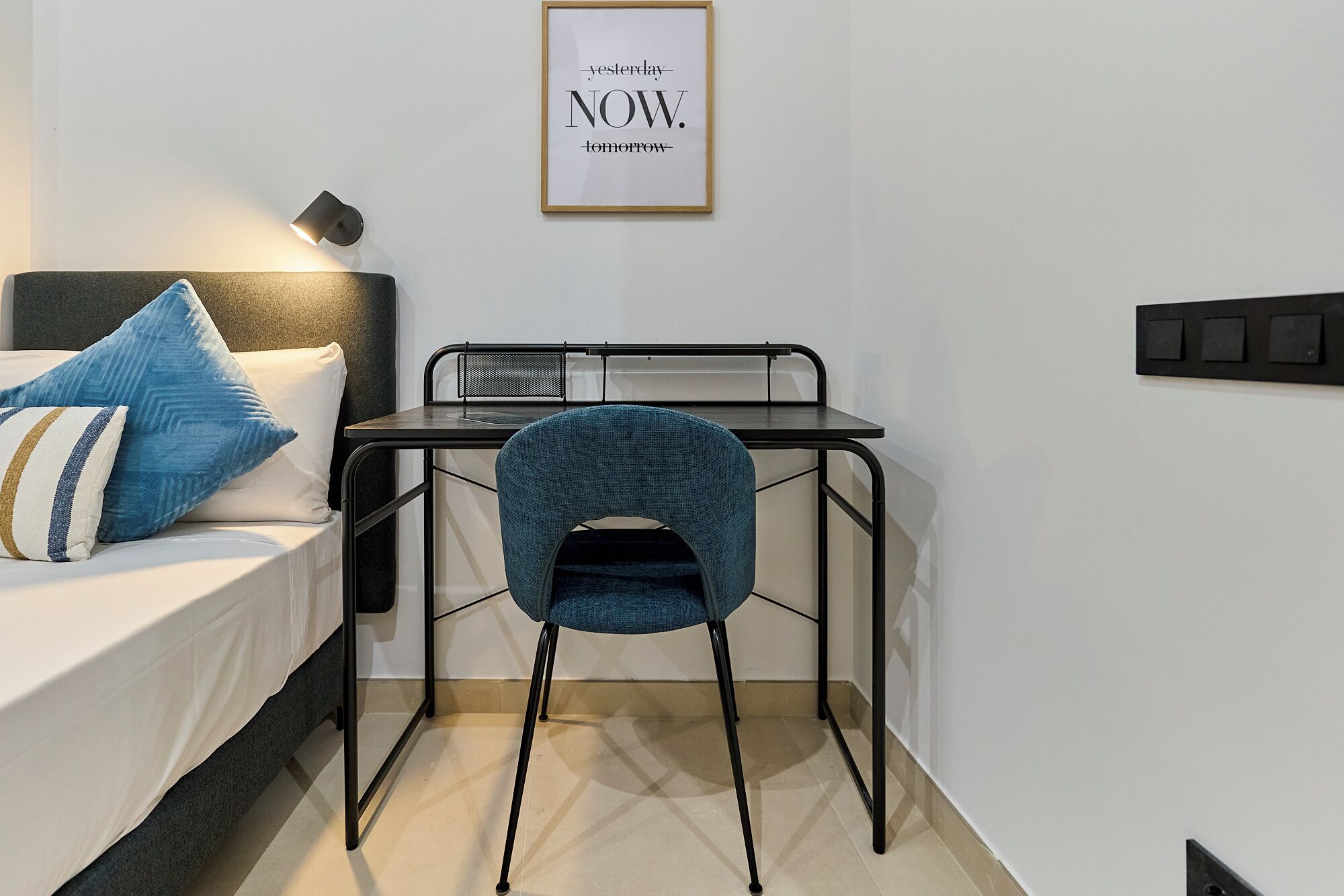 A small, modern room with a bed, a desk, and a chair. There is a framed quote on the wall.