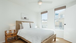 Un dormitorio con cama de madera, ropa de cama blanca, una mesita de noche con lámpara y una ventana con vistas a edificios.