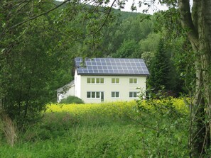 Ett hus med solpaneler, omgivet av frodig grönska och ett fält med gula blommor.