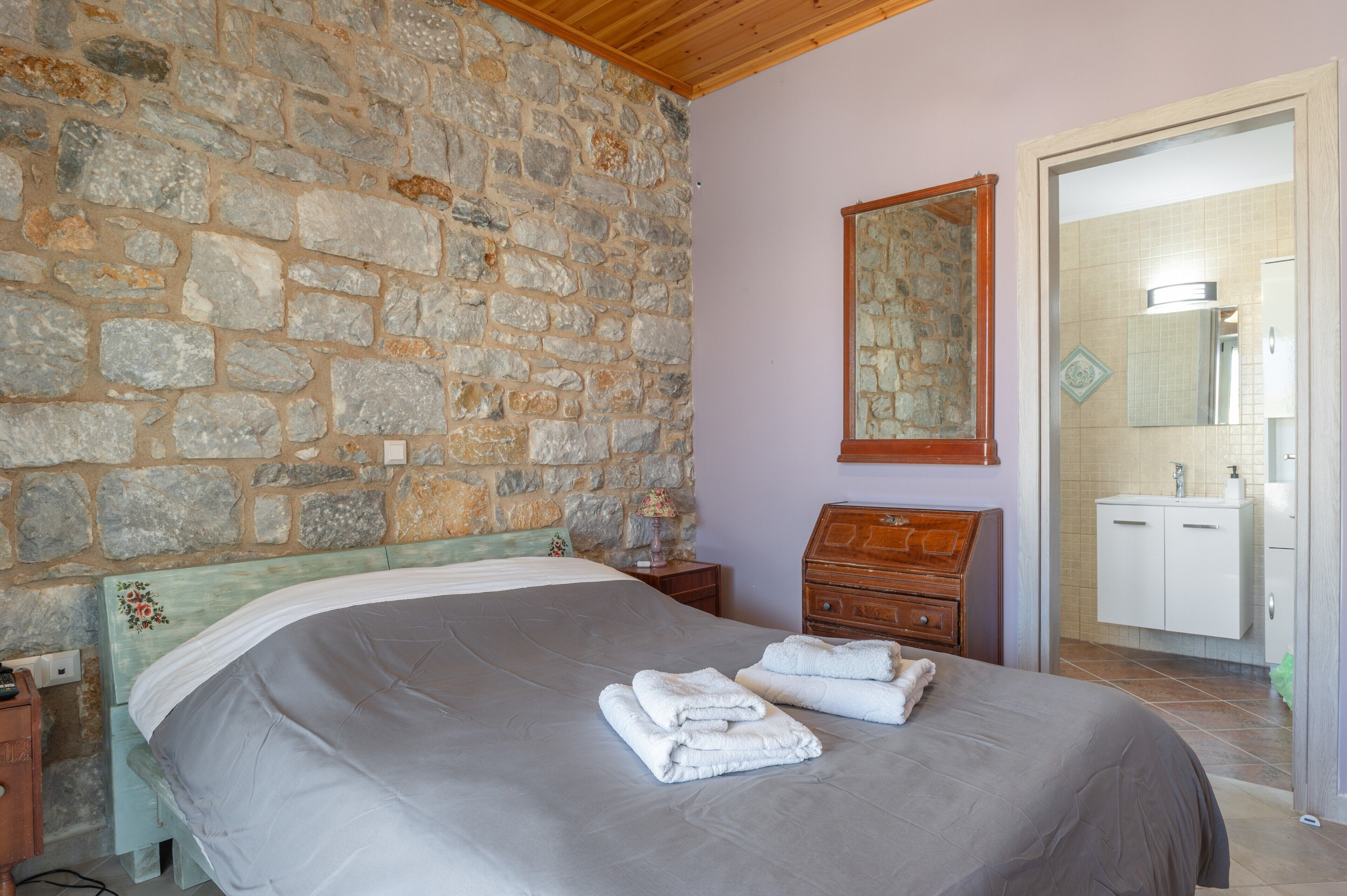 A bedroom with a stone wall, a bed with a grey bedspread, two folded white towels, a wooden nightstand, a wooden dresser, and a bathroom visible through an open door.