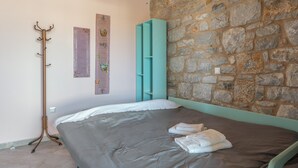 Une chambre avec un mur en pierre, un lit avec une literie grise, une armoire turquoise et un porte-manteau.