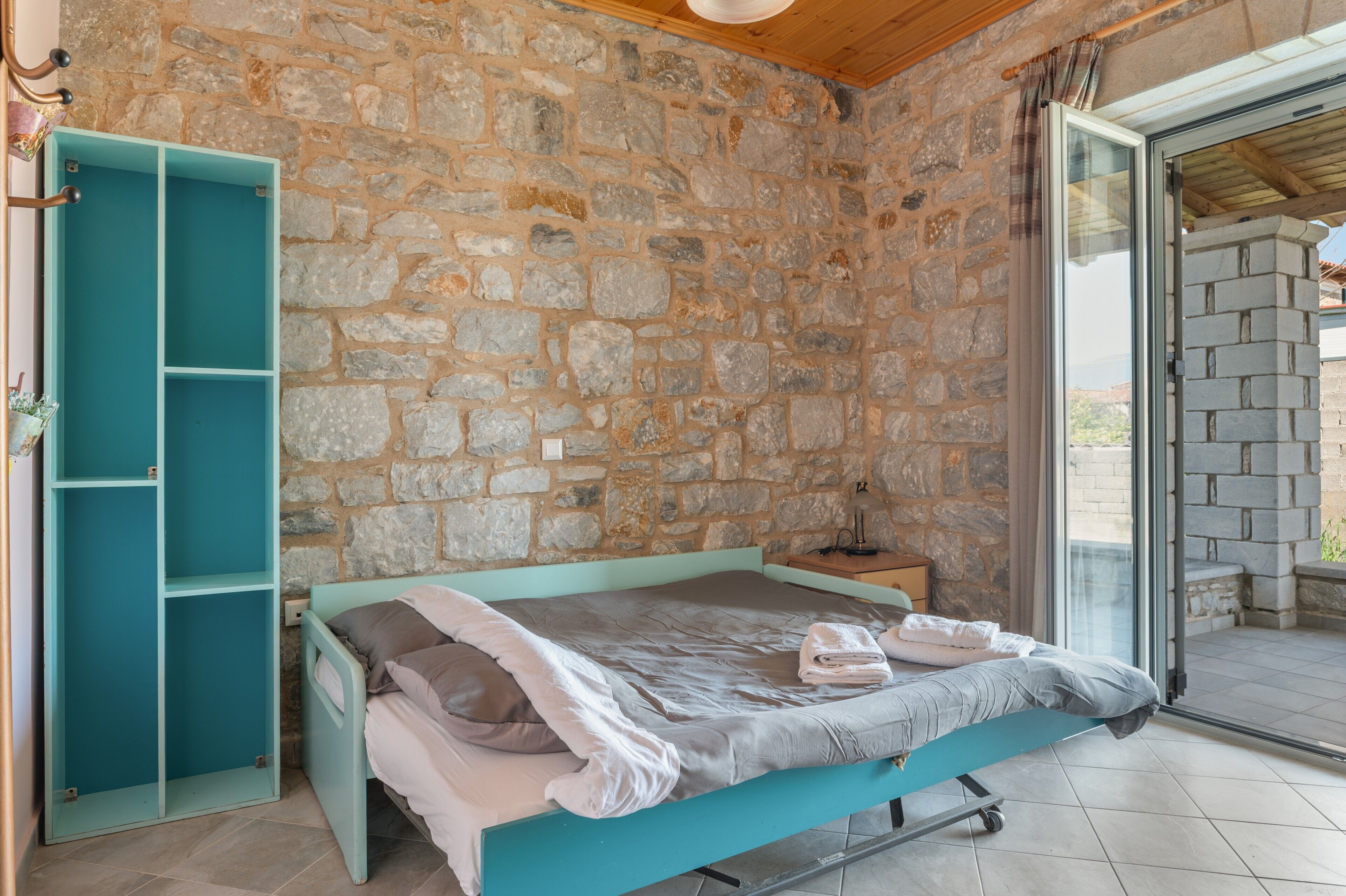 A room with a stone wall, a bed with a teal frame, a small table, and a sliding glass door leading to an outdoor area.