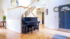 A spacious living room with a grand piano, wooden staircase, and a blue door.