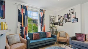 Un salón con un sofá azul, dos sillones, una mesa de centro y una ventana con cortinas coloridas.