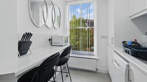 Una cocina moderna con encimera blanca, dos taburetes negros, una ventana con vistas a zonas verdes y un electrodoméstico blanco.