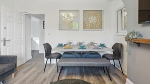 Un comedor con una mesa dispuesta para cuatro personas, dos obras de arte en la pared, un televisor montado y un banco gris.