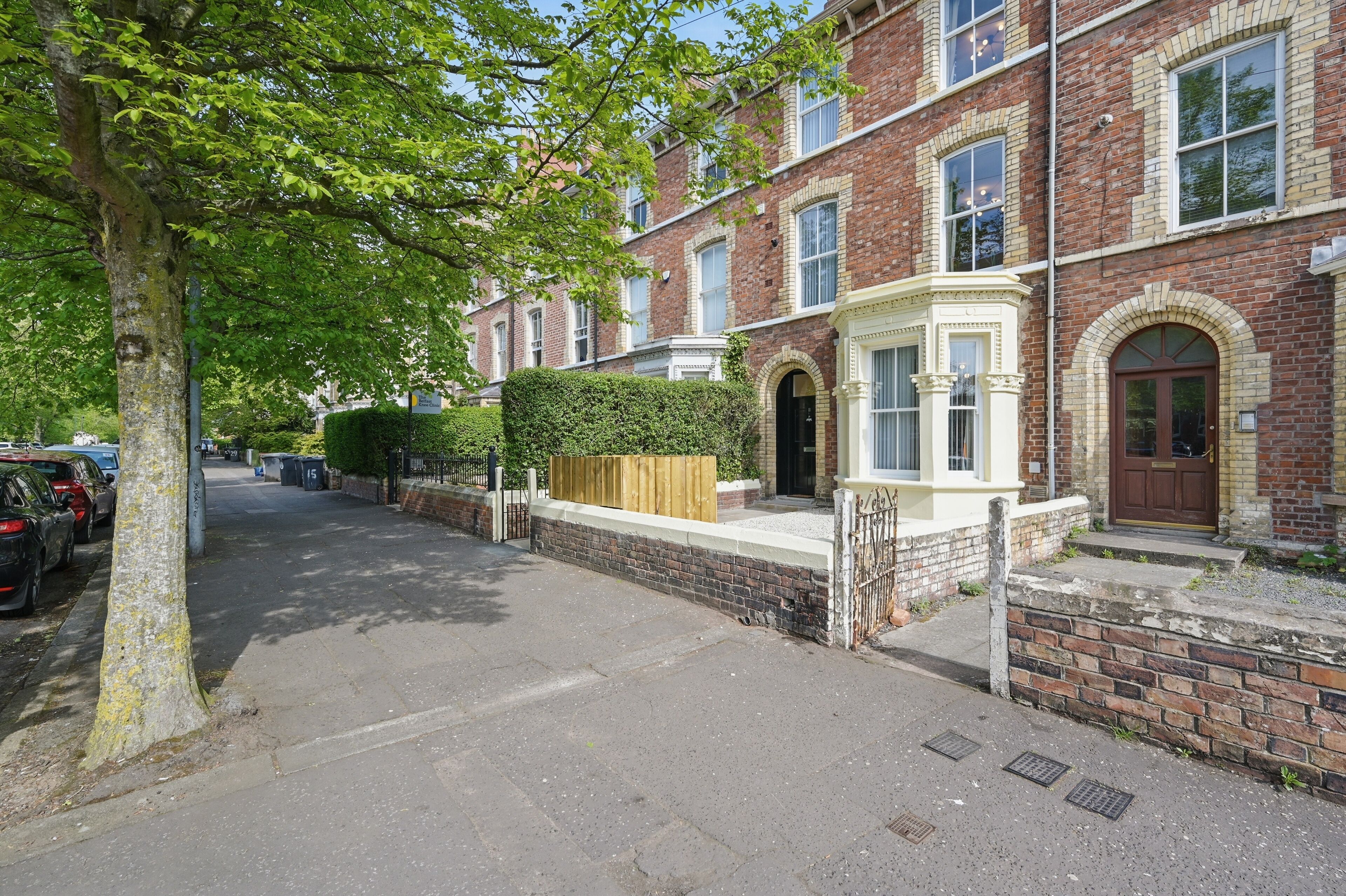 A residential street with brick houses, a tree-lined sidewalk, and parked cars.