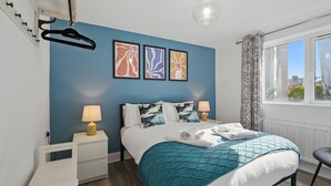 Un dormitorio con una cama, dos mesitas de noche, una ventana con cortinas y tres cuadros en la pared.