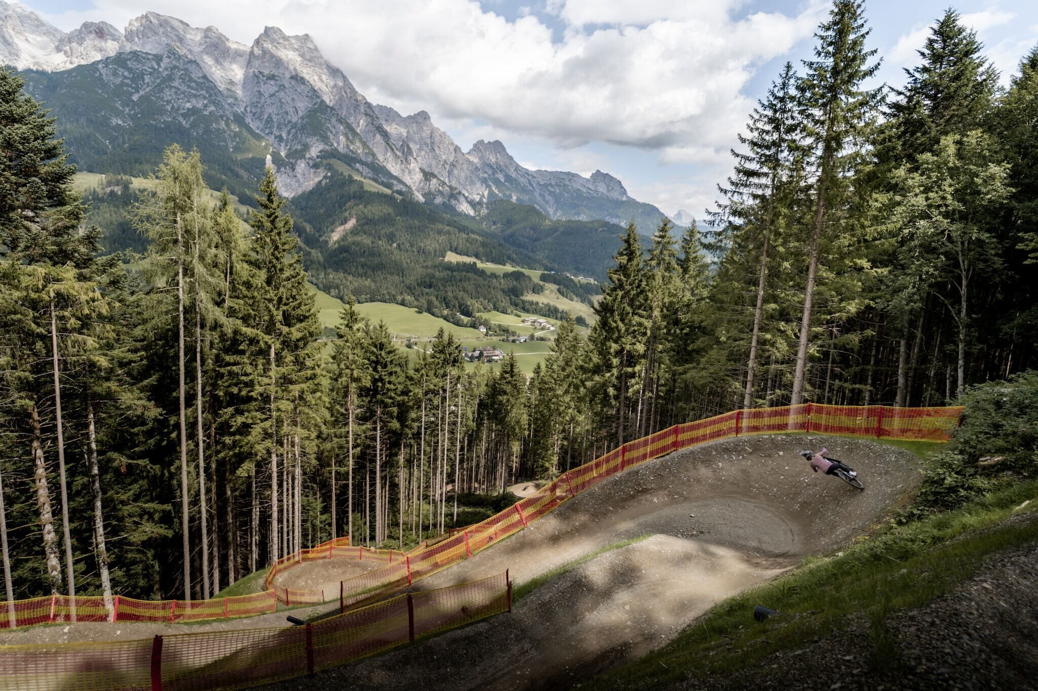 A mountain biking track with a rider, surrounded by tall trees and mountains in the background.