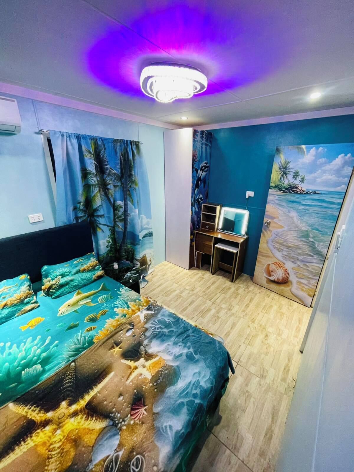 Un dormitorio con una cama decorada con un mural tropical, un escritorio con una computadora y un cuadro con motivos playeros en la pared.