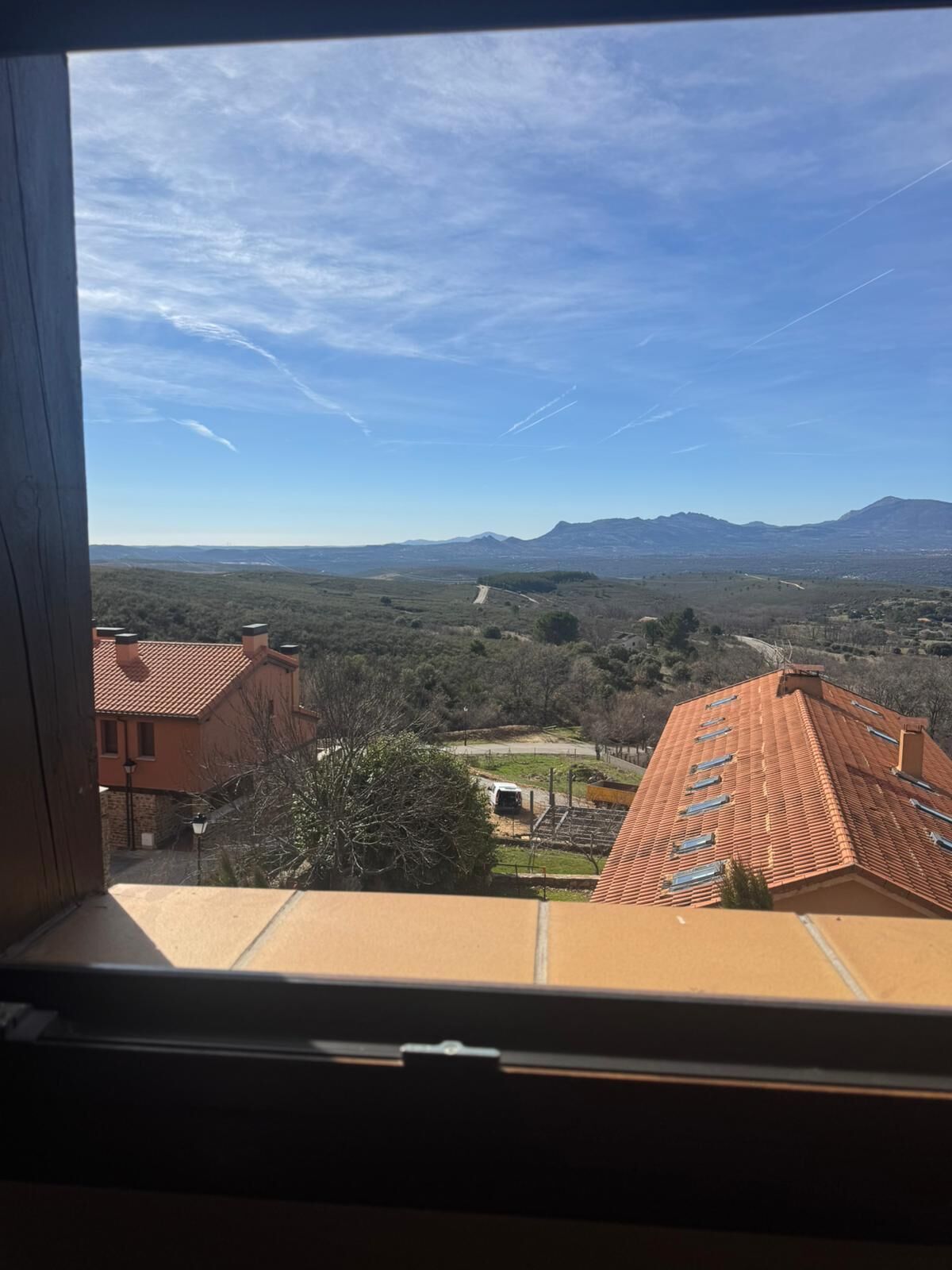Vista des d'una finestra sobre un paisatge rural amb muntanyes al capvespre, un cel clar i un edifici amb teulada vermella.