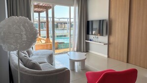 Et moderne hotelværelse med en grå sofa, en rød lænestol, et fjernsyn på en hylde og udsigt til en pool og udendørsområde.