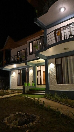 A two-story house with a balcony, a small porch with steps, and a stone fire pit in the yard. - Sylvan Woods Cottage (Manali)