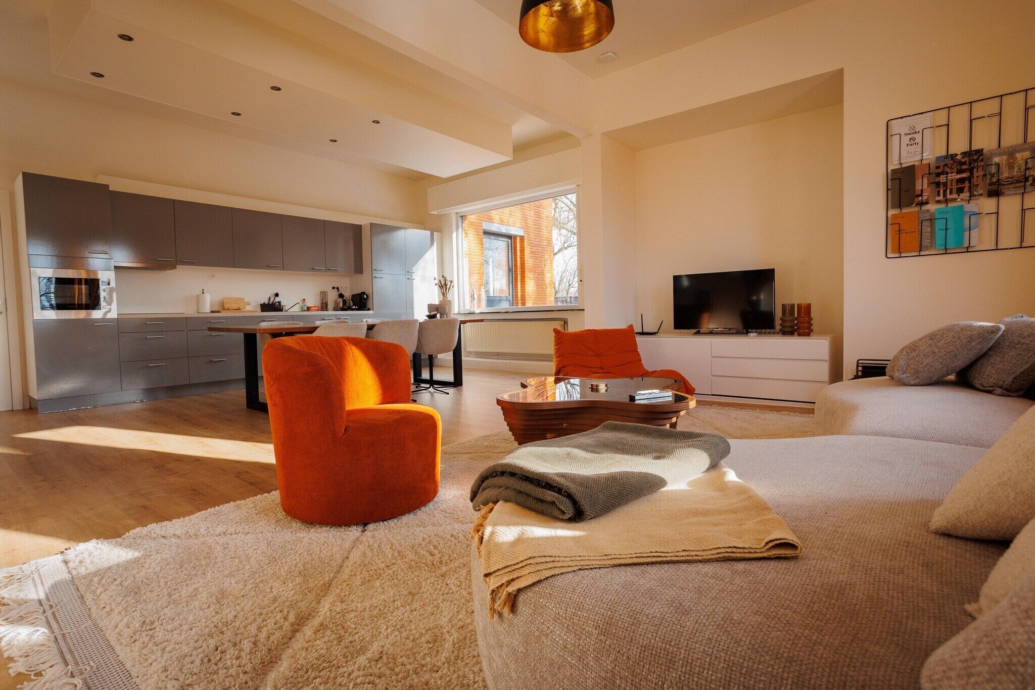 A modern living room with a sofa, a coffee table, and a TV. There is a kitchen area with cabinets and a window.