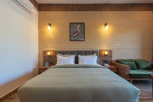 Habitación de hotel con una cama grande, dos mesitas de noche con lámparas, un sillón verde y un cuadro enmarcado en la pared.