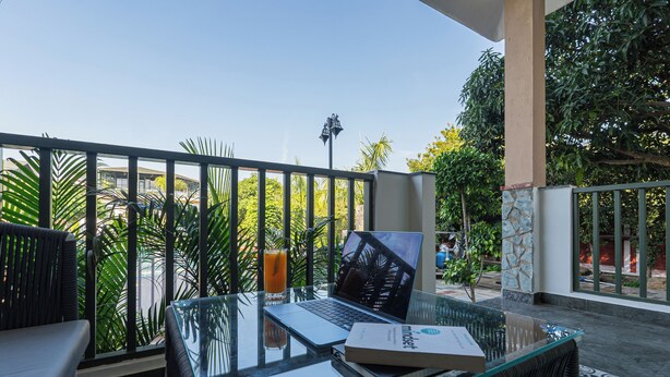 A glass-top table with a laptop, a book, and a glass of orange juice on a balcony with a view of greenery and a building in the background. - Trideus The Wild Fraser Corbett (Kaladhungi)