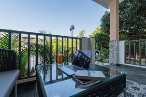 Uma mesa com tampo de vidro, um laptop, um livro e um copo de suco de laranja numa varanda com vista para o verde e um edifício ao fundo.