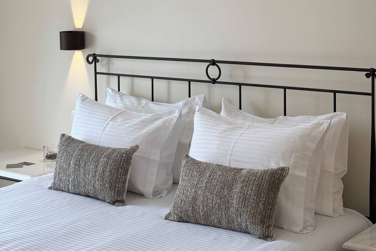 Una cama bien hecha con almohadas blancas y grises, un cabecero de metal negro y una lámpara de pared.