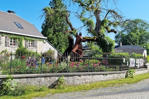 Ein Steinhaus mit geneigtem Dach, ein umzäunter Garten mit bunten Blumen und ein Baum mit einer Pferde Skulptur.