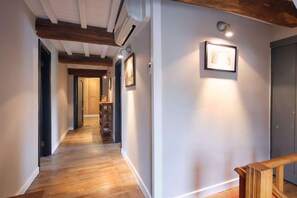 Een hal met houten vloer, witte muren en een plafond met zichtbare balken. Er hangen afbeeldingen in een lijst aan de muur en bevindt zich een deur aan de rechterkant.